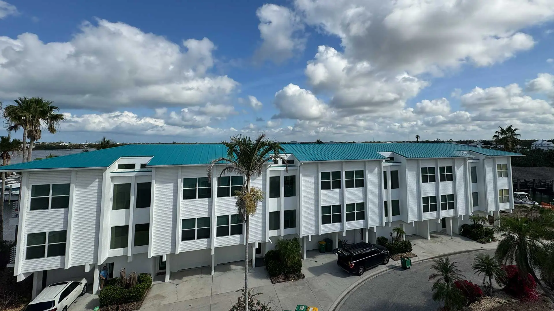 elevated, wide-angle view of a multi-unit white residential building with a bright teal metal roof under a partly cloudy blue sky, flanked by palm trees
