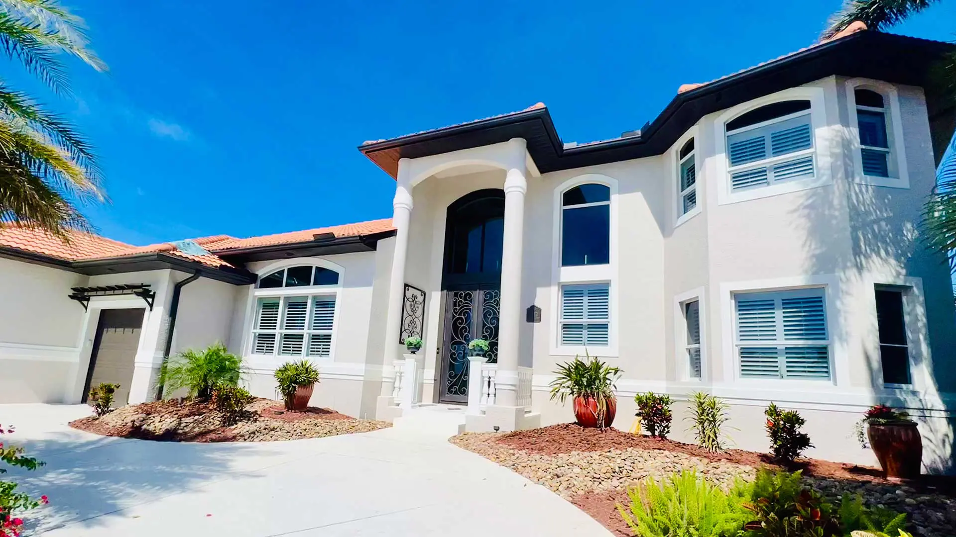 front entrance of a large two-story mediterranean-style home, featuring a grand arched entryway with white columns, a dark ornamental wrought iron double front door, a concrete driveway, terracotta tile roofing, and landscaped garden beds under a bright blue sky