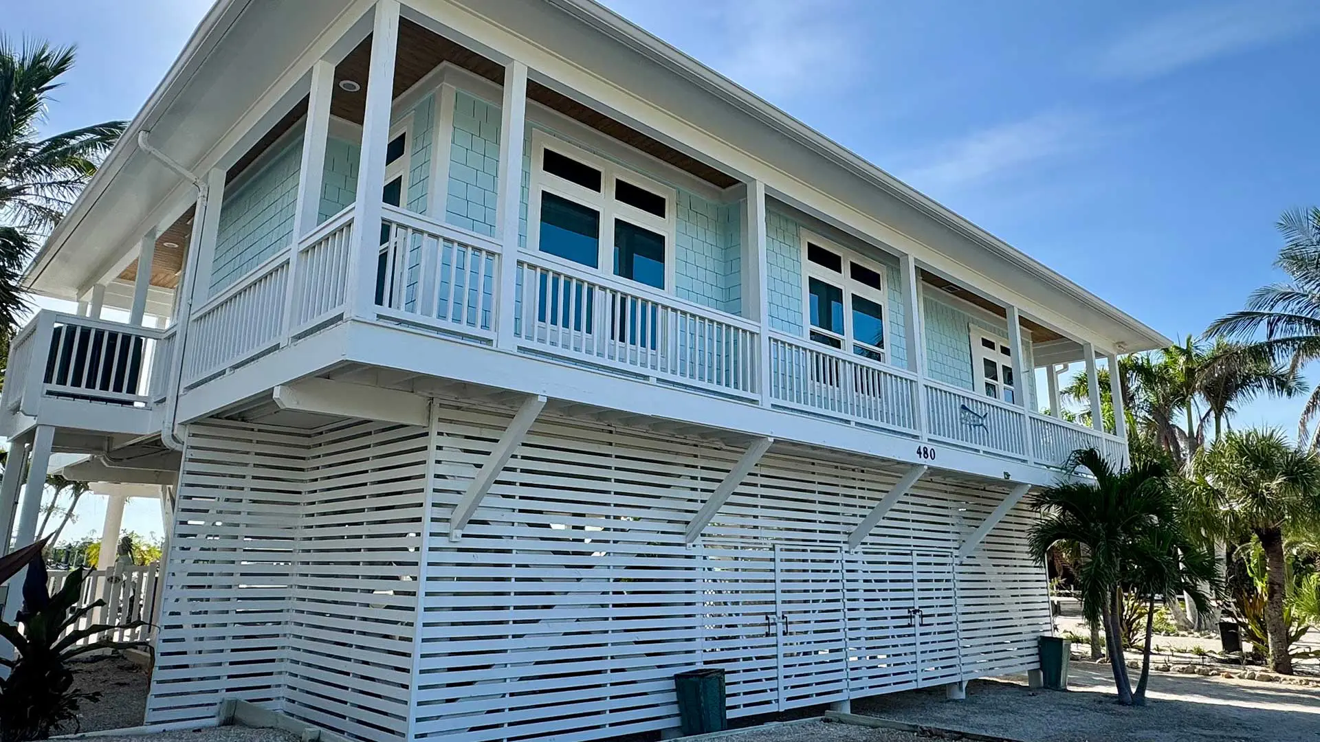 a two-story coastal home in Southwest Florida, featuring light blue shake siding and white trim, elevated on white horizontal lattice stilts with palm trees against a bright blue sky