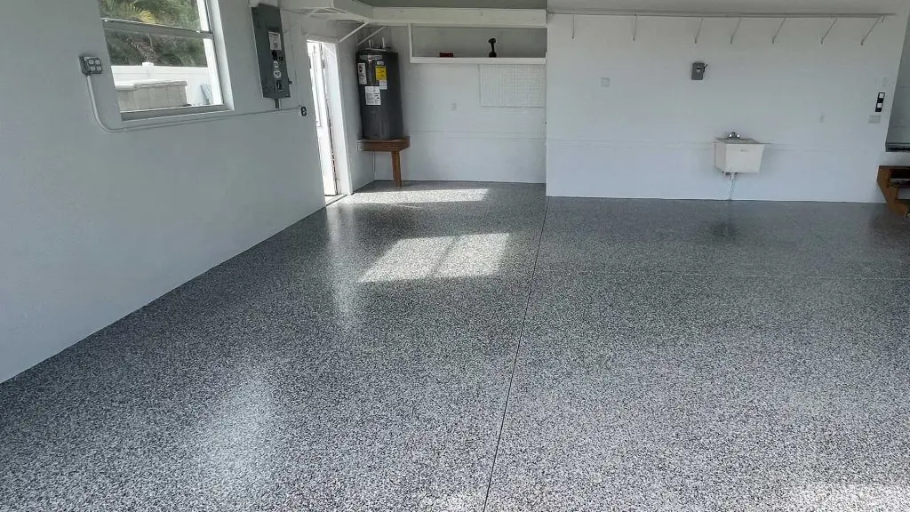 interior view of an empty garage featuring a freshly finished gray speckled epoxy floor, white walls, a water heater in the corner, a small utility sink mounted on the wall, and natural light streaming in from a side door and window