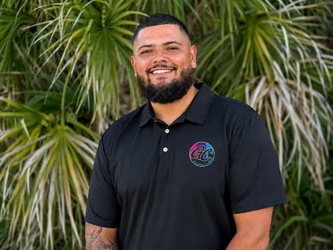 Portrait of Andrew Curtis smiling in front of a green palm leaf background, wearing a black Gulf Coast Painting & More polo shirt