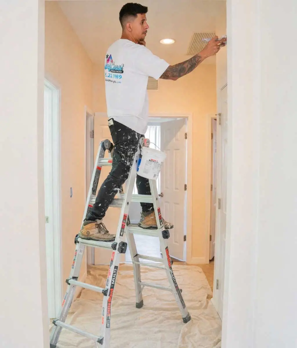professional painter standing on an aluminum stepladder while painting a doorway trim inside a home hallway, wearing a white t-shirt and paint-splattered black pants with a drop cloth protecting the floor below