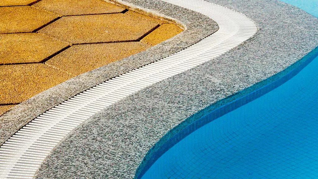 close-up view of a swimming pool edge, featuring bright blue water, a gray granite pool coping with a white plastic overflow drainage grate, and a textured tan interlocking paver pool deck
