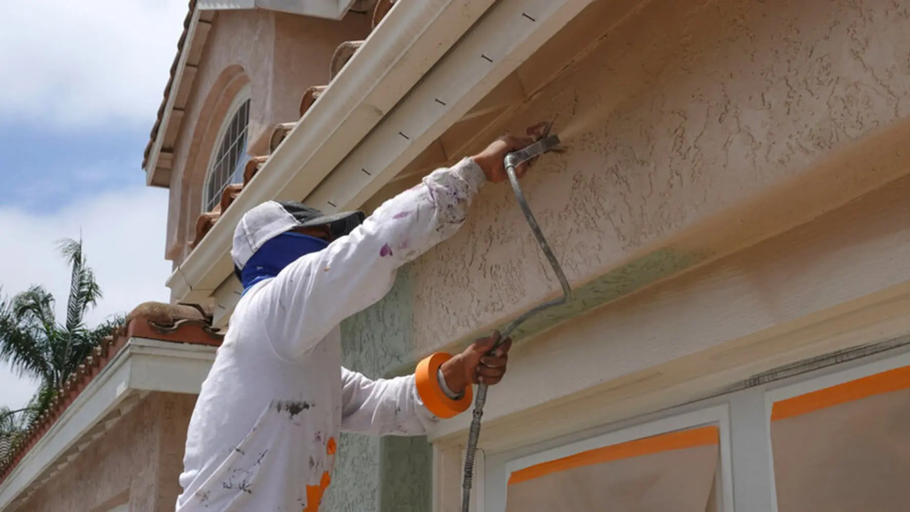 A professional painter in white coveralls spraying the stucco exterior of a Florida home with a paint sprayer.