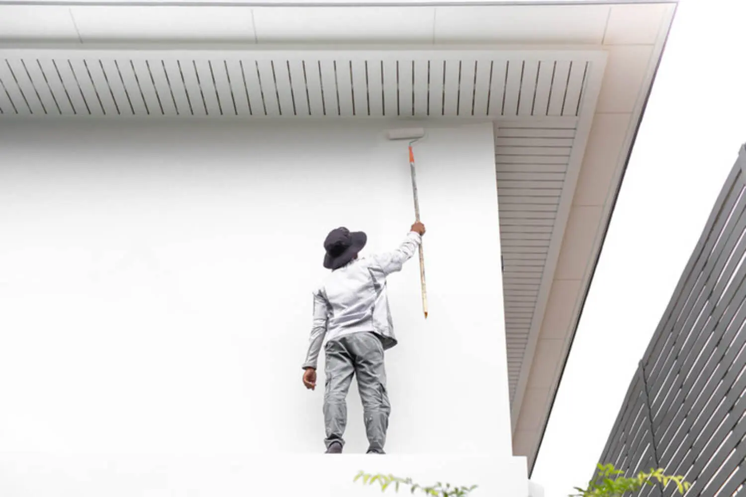 A professional painter in work clothes rolling white paint on the exterior wall of a residential home.