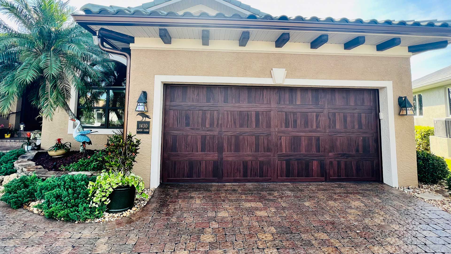 front view of a single-story home's garage, featuring a dark faux-wood finish double garage door, beige stucco walls, a brick paver driveway, and a tiled roof with dark wood rafter tails next to a palm tree and landscaped garden bed