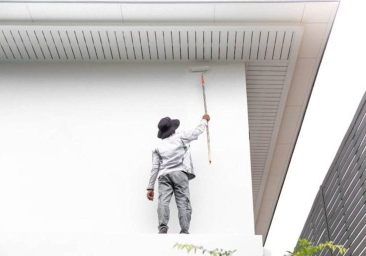 A professional painter in work clothes rolling white paint on the exterior wall of a residential home.