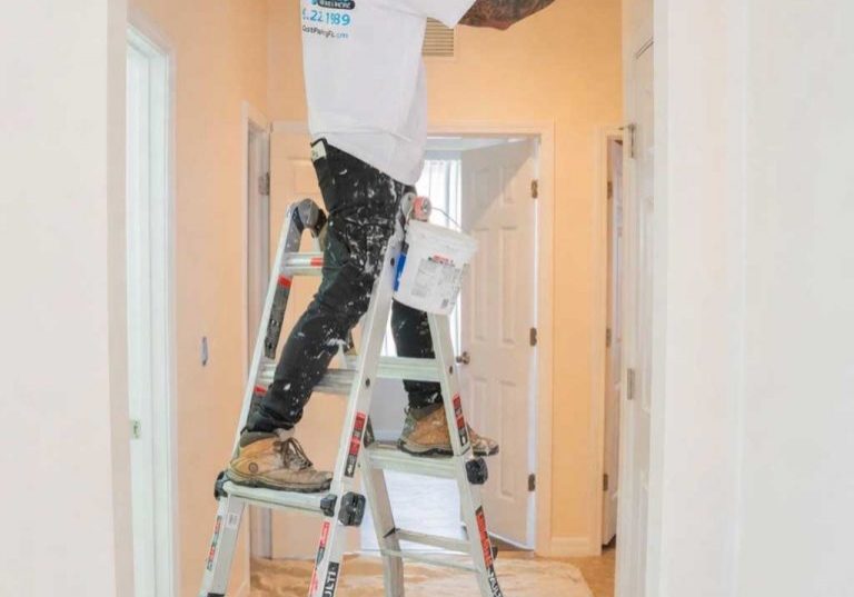 professional painter standing on an aluminum stepladder while painting a doorway trim inside a home hallway, wearing a white t-shirt and paint-splattered black pants with a drop cloth protecting the floor below