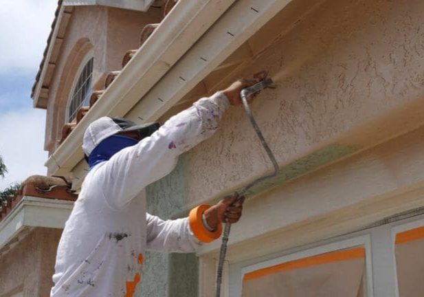 A professional painter in white coveralls spraying the stucco exterior of a Florida home with a paint sprayer.